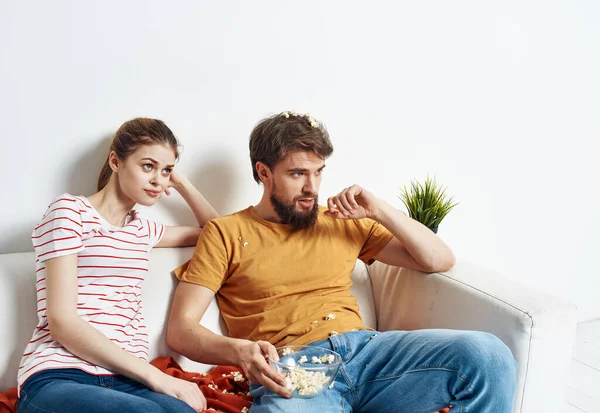 A man with popcorn in a plate and a woman are sitting on the couch watching TV and watching movies