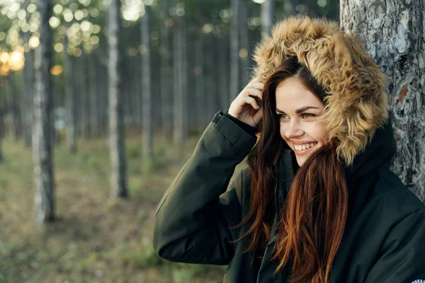 A woman in an autumn jacket with a hood near a tree on a forest background — Stock Photo, Image