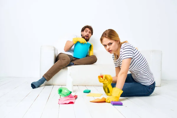 A man sits on a sofa with a bucket of a woman on the floor with a detergent for kitchen interior chores — Stock Photo, Image