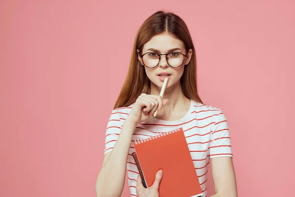 Estudante do sexo feminino com blocos de notas em fundo rosa gestos com as mãos óculos em seu rosto listrado t-shirt modelo — Fotografia de Stock