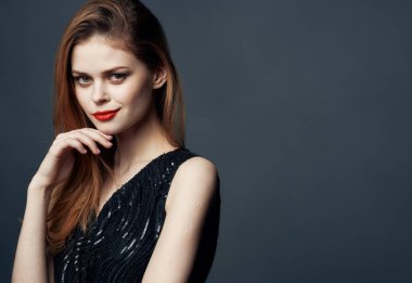 Siyah elbiseli, gri arka planda kızıl saçlı bir kadın elleriyle işaret ediyor.