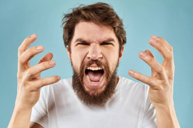 Beyaz tişörtlü sakallı adam. Duyguları, elleri ve yüz ifadesinden memnun olmayan mavi arka plan.