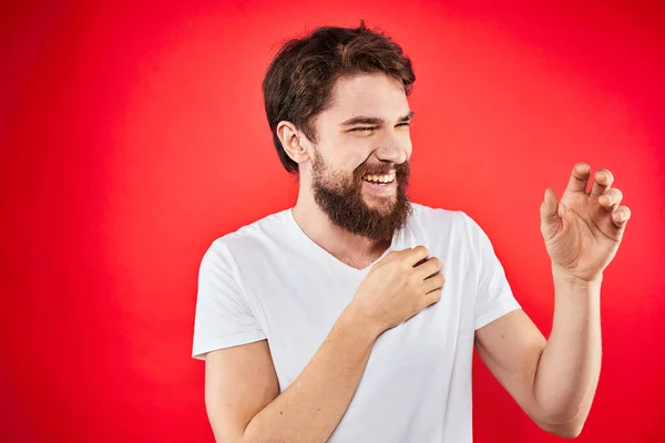 A man in a white T-shirt with a beard gestures with his hands emotions red background