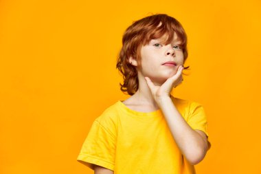 Kızıl saçlı bir çocuk elini yüzüne yakın tutuyor ve ileriye bakıyor.