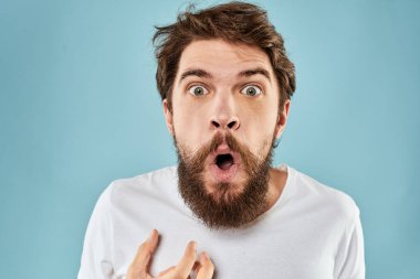 Huysuz yüz ifadesi olan bir adam. Stüdyo tarzı mavi arka planda el kol hareketi yapıyor.