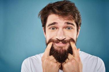 Sakallı adam, yüz ifadesinden rahatsız. Duygular, yakın plan mavi arka plan, beyaz tişört.