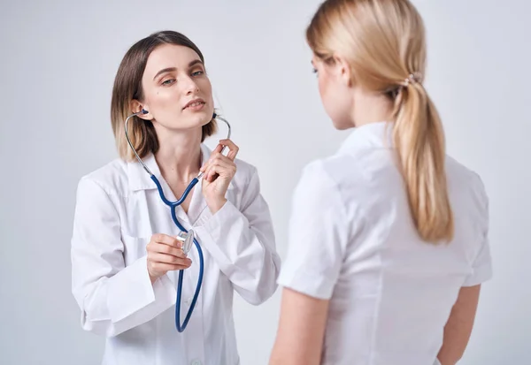 Médico com um estetoscópio examina uma mulher em uma camiseta branca em um fundo leve — Fotografia de Stock