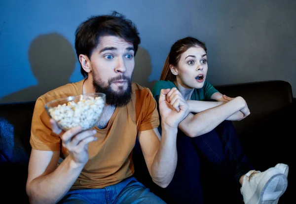 a man and a frightened woman watching a movie in the evening on the couch with a plate of popcor