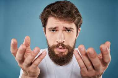 Sakallı adam, yüz ifadesinden rahatsız. Duygular, yakın plan mavi arka plan, beyaz tişört.
