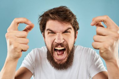 Beyaz tişörtlü sakallı adam. Duyguları, elleri ve yüz ifadesinden memnun olmayan mavi arka plan.