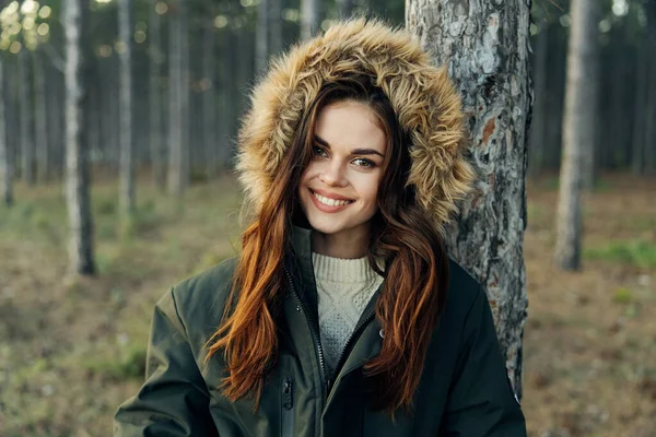 Mujer en chaqueta caliente con una capucha cerca de un árbol en el bosque sonrisa mirada atractiva —  Fotos de Stock