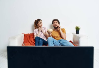 Arkadaşlar, erkek ve kadın kanepede oturur, televizyonun önünde ve saksıda bir çiçek.