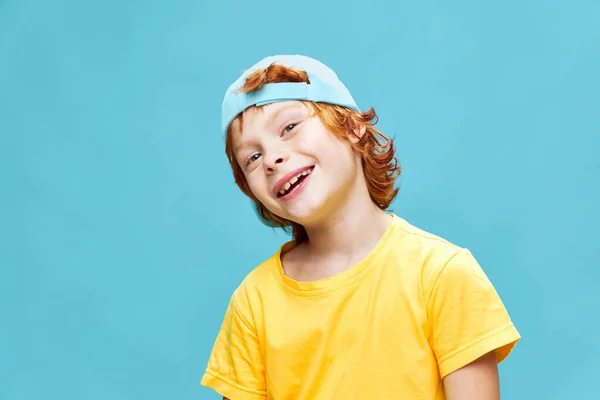 Усміхаючись широко рудий хлопчик в жовтій футболці з шапочкою на голові синій фон — стокове фото