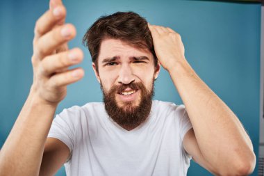 Sakallı adam, yüz ifadesinden rahatsız. Duygular, yakın plan mavi arka plan, beyaz tişört.