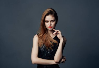 Siyah elbiseli, gri arka planda kızıl saçlı bir kadın elleriyle işaret ediyor.