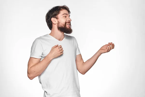 Homem barbudo em gesto t-shirt branco com as mãos emoções luz fundo — Fotografia de Stock