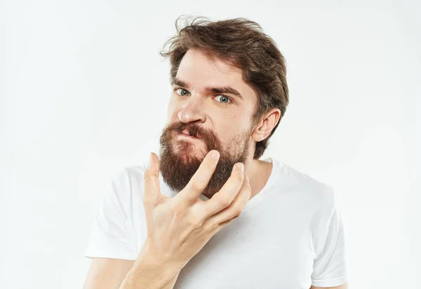 Un hombre desconcertado con barba y con una camiseta blanca sobre un fondo claro — Foto de Stock