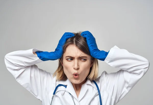 Emotional woman in blue medical gloves touches her head with her hands on a light background