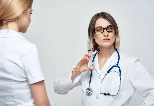 Медсестра держит шприц в руке на светлом фоне со стетоскопом и пациенткой — стоковое фото