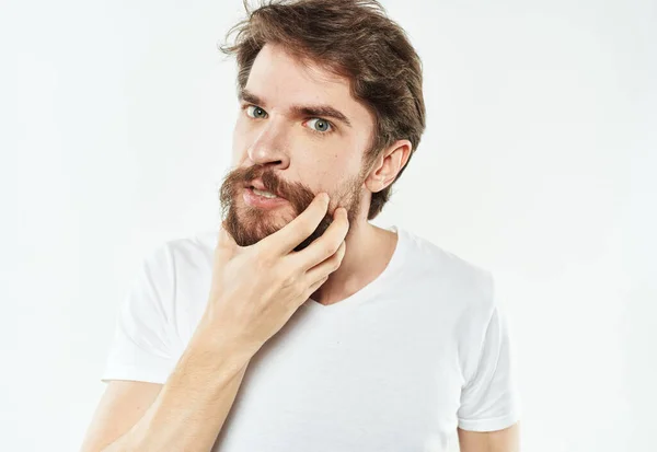 Um homem perplexo com barba e uma t-shirt branca sobre um fundo claro — Fotografia de Stock