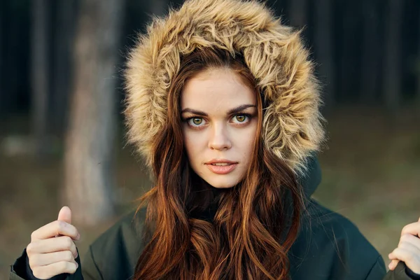 Uma mulher em um casaco com um capuz olha para a frente contra o fundo da floresta — Fotografia de Stock