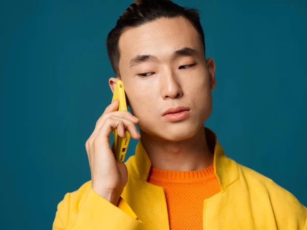 Emotionaler asiatischer Mann auf blauem Hintergrund telefoniert mit gelber Jacke — Stockfoto