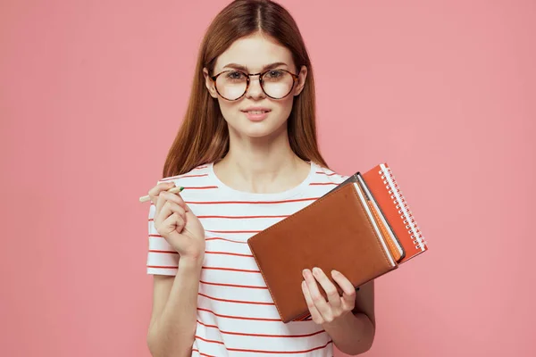 Female student with notepads on pink background gesturing with her hands glasses on her face striped t-shirt model — Stock Photo, Image