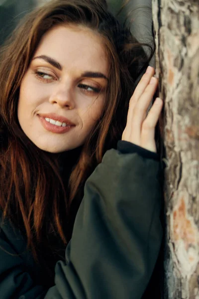 Mujer en una chaqueta apoyada en un árbol vista lateral sonrisa viaje —  Fotos de Stock