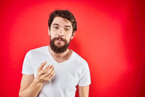 A man in a white T-shirt with a beard gestures with his hands emotions red background