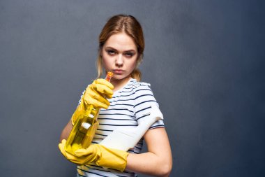 Duygusal temizlikçi kadın sarı lastik eldiven ve deterjan gri arka plan giyiyor.