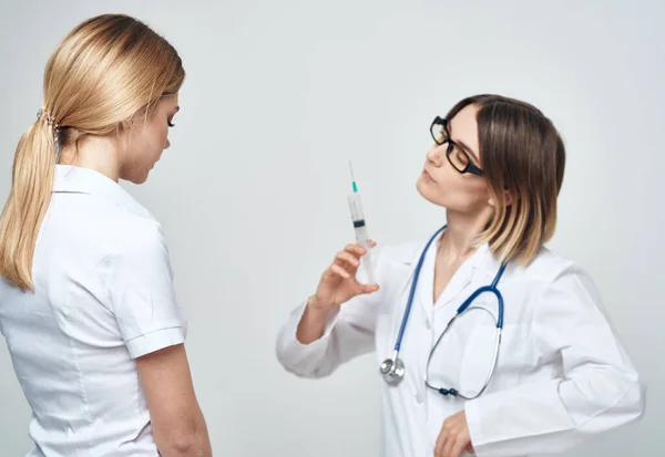 Una donna infermiera tiene in mano una siringa su uno sfondo chiaro con uno stetoscopio e una paziente — Foto Stock