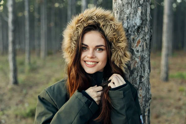 Glimlachende vrouw in warme jas met een kap in de buurt van een boom op een bosachtergrond bijgesneden uitzicht — Stockfoto