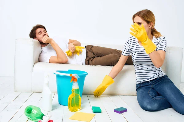 Un hombre yace en un sofá. Una mujer limpia la limpieza del hogar — Foto de Stock