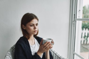 Kadının biri elinde bir fincan kahveyle pencerenin yanındaki ekosenin arkasına saklanıyor. 