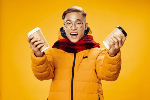 A man in a yellow winter jacket warming drink in his hands cool checkered scarf