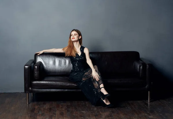 Uma mulher em um vestido preto senta-se em um sofá de couro em uma sala cinza em pleno crescimento — Fotografia de Stock