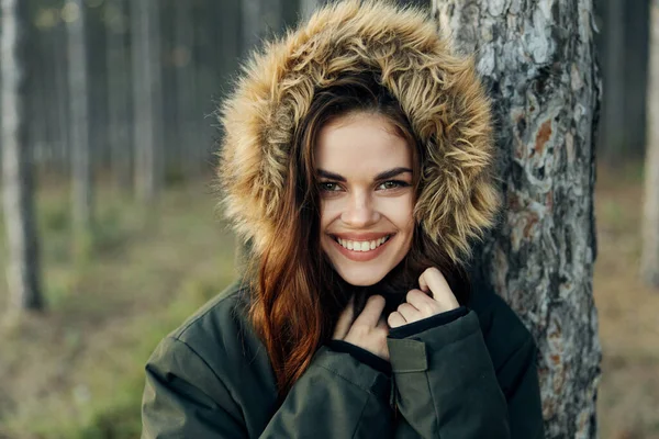 Mujer sonriente en una chaqueta con una capucha cerca de un árbol en la naturaleza —  Fotos de Stock
