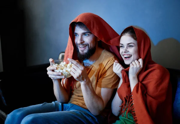 Ένας άντρας με ένα πιάτο ποπ κορν και μια συναισθηματική γυναίκα κάτω από μια κόκκινη κουβέρτα στον καναπέ. — Φωτογραφία Αρχείου