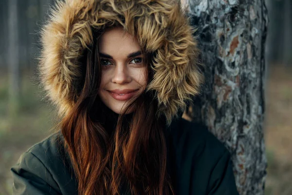 Una mujer con una chaqueta caliente con una capucha cerca de un árbol sobre un fondo forestal —  Fotos de Stock