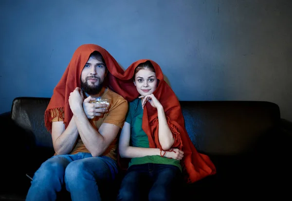 Una mujer y un hombre bajo una manta roja en el sofá viendo la televisión por la noche — Foto de Stock