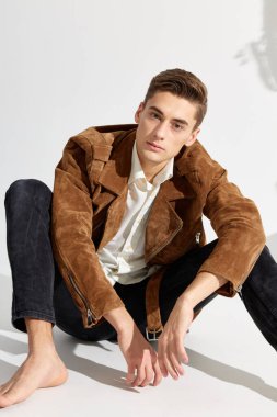 Kahverengi ceketli ve siyah pantolonlu zarif bir adam parlak bir odada yerde oturuyor.