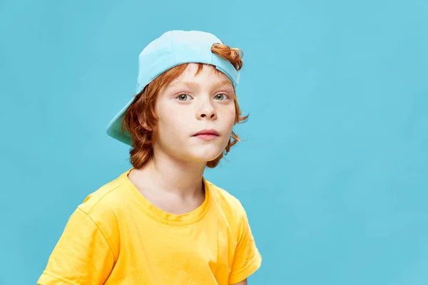 一个好奇的红头发男孩，头上戴着一顶蓝色的帽子，期待着黄色的T恤 — 图库照片