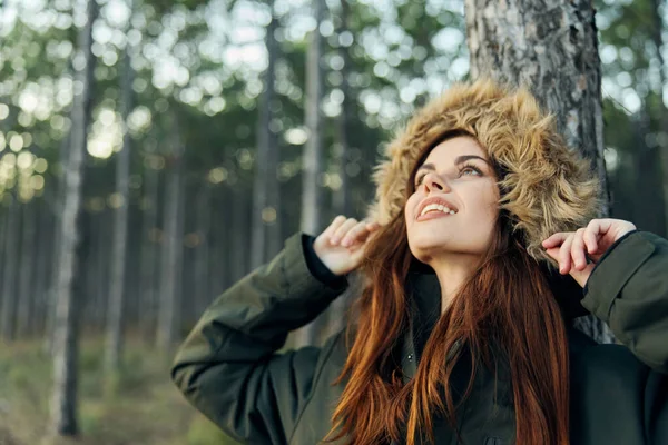 Una mujer con una chaqueta natural mira hacia arriba en el bosque contra el fondo de los árboles —  Fotos de Stock