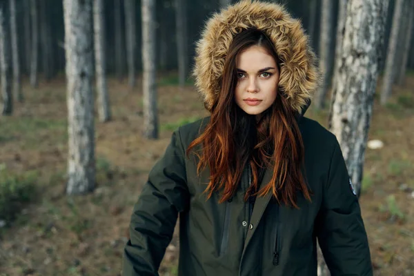 Mujer en una chaqueta negra con una capucha en la naturaleza mira hacia adelante sobre el fondo del bosque —  Fotos de Stock