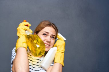 Duygusal temizlikçi kadın sarı lastik eldiven ve deterjan gri arka plan giyiyor.