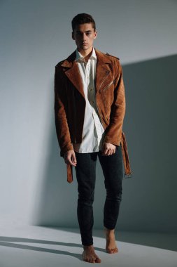 Düğmeleri iliklenmemiş ceketli ve pantolonlu genç bir adam ve gri arka planda tamamen büyümüş bir gömlek.