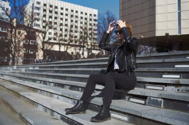 Deri ceketli bir kadın, tiyatronun yanındaki merdivenlerde oturuyor.