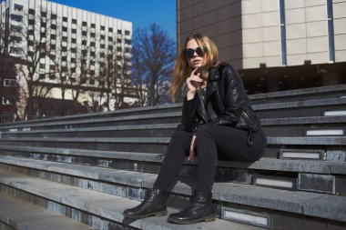 Deri ceketli kadın, sonbaharda caddede binanın yanındaki merdivenlerde oturuyor.