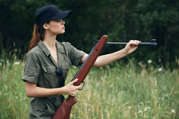 녹색 작업복을 입은 채 야외에서 자연 무기를 배경으로 총을 장전하고 있는 여자 — 스톡 사진