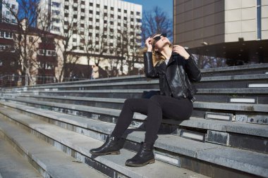 Deri ceketli kadın, sonbaharda caddede binanın yanındaki merdivenlerde oturuyor.